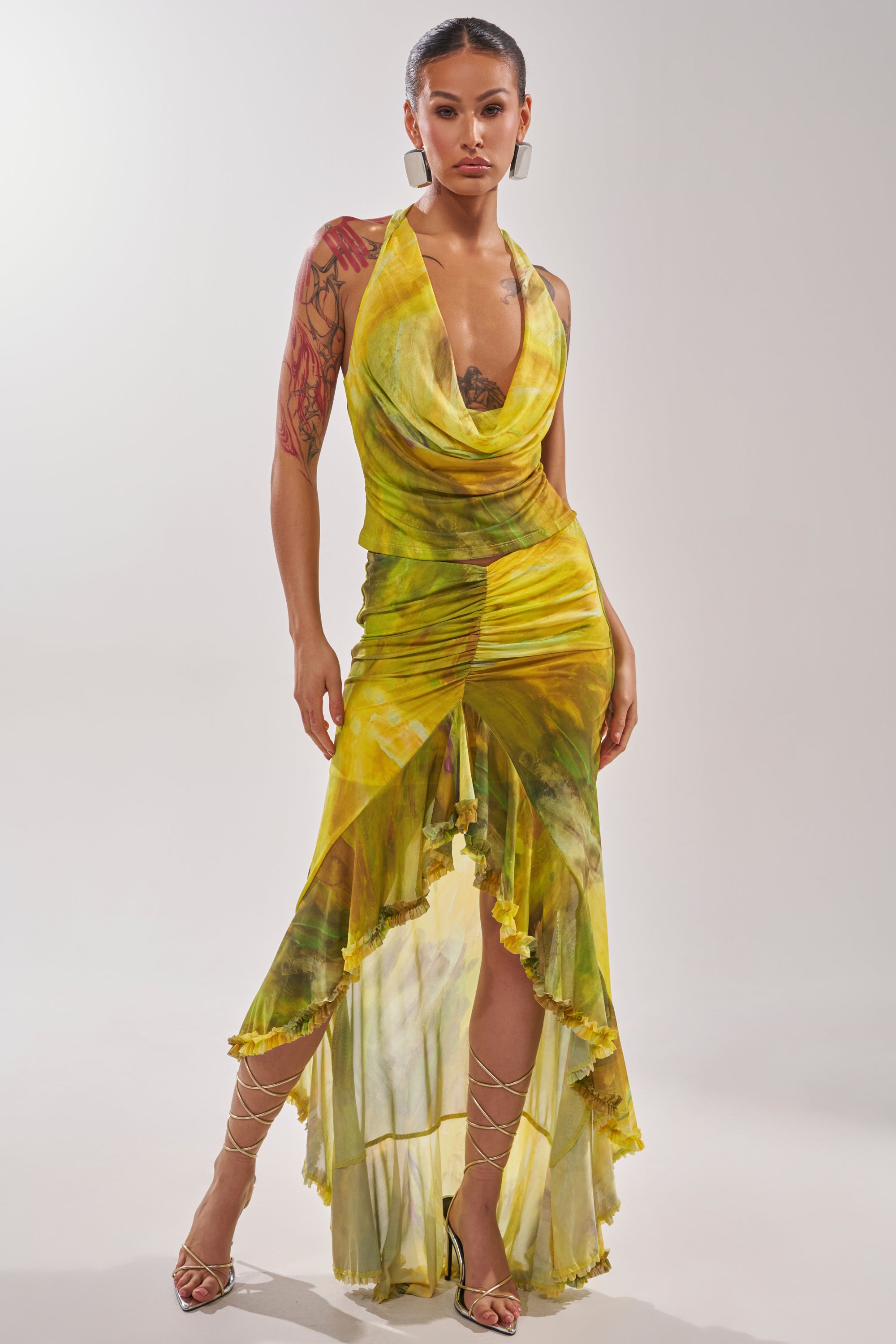 A woman stands confidently in the TELL ME MORE HIGH LOW SKIRT IN YELLOW, styled with strappy sandals, hoop earrings, and a visible arm tattoo. Her yellow-green tie-dye halter dress features an asymmetrical high-low skirt and draped neckline against a plain background. 0