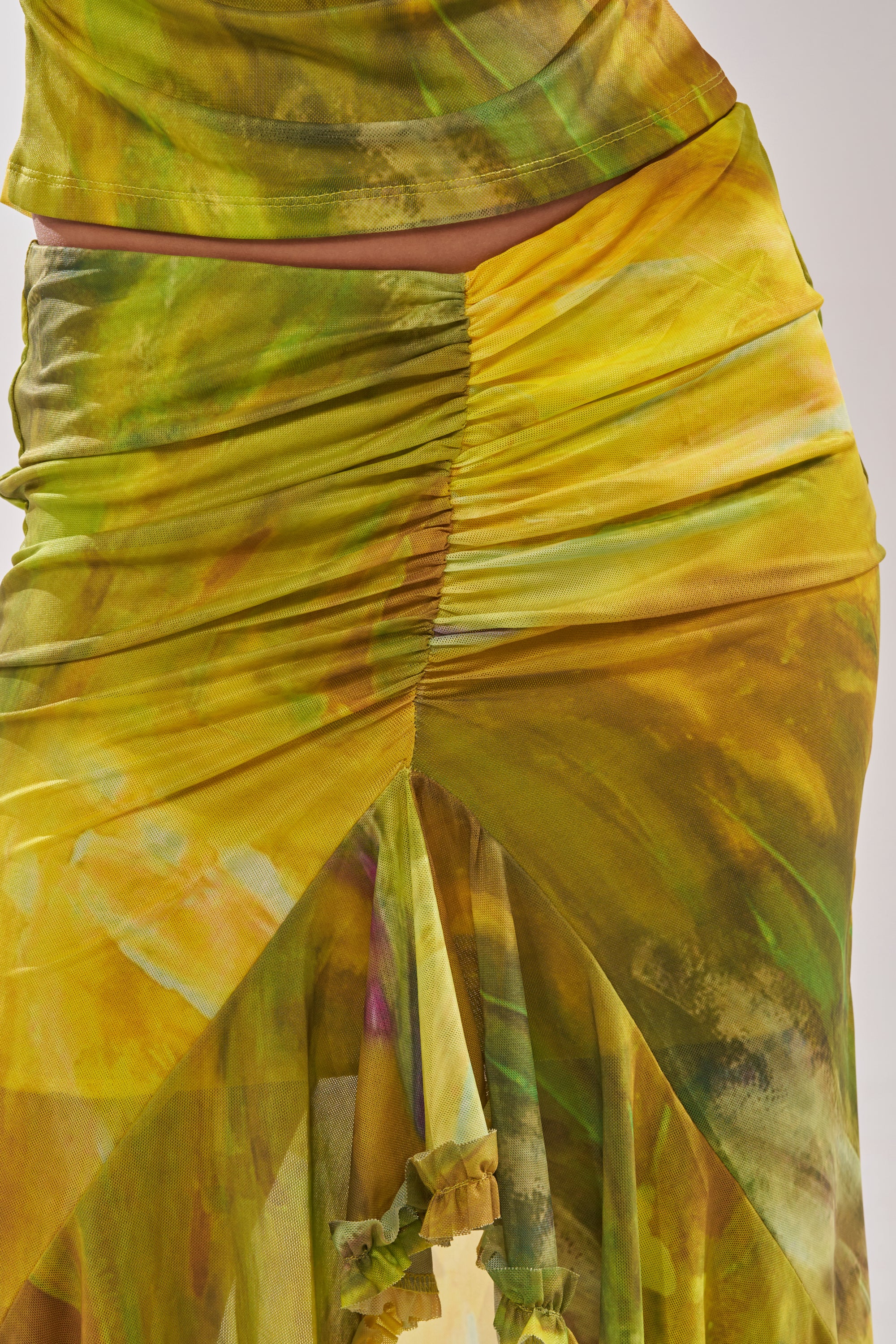 Close-up of the TELL ME MORE HIGH LOW SKIRT IN YELLOW, showcasing yellow and green tie-dye patterns, a ruched waist, gathered fabric, and draping details around the waist and upper thigh. 9
