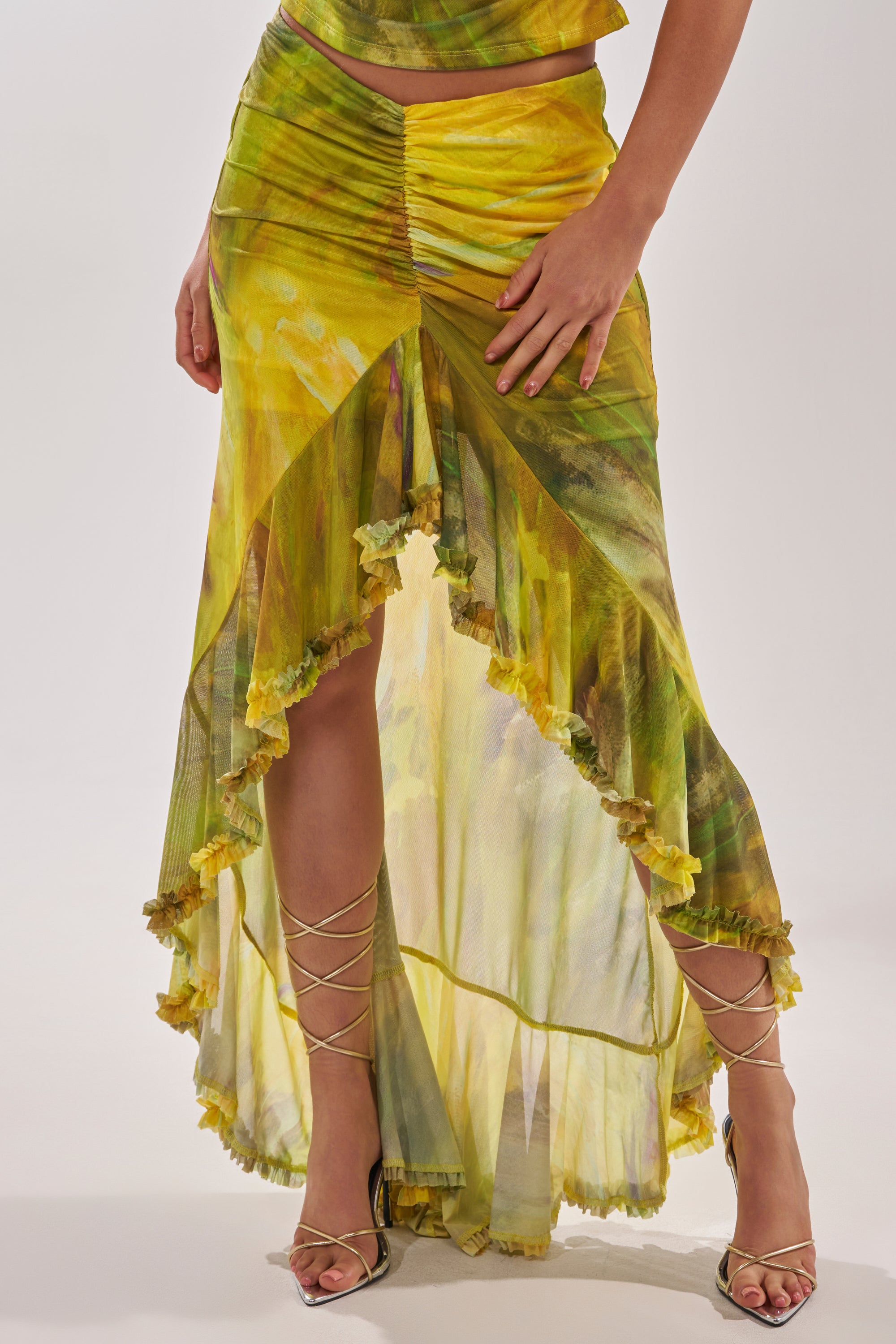 A person models the TELL ME MORE HIGH LOW SKIRT IN YELLOW—featuring a ruffled high-low hem and ruched details—paired with strappy heels. Only their lower body is shown against a plain background. 1