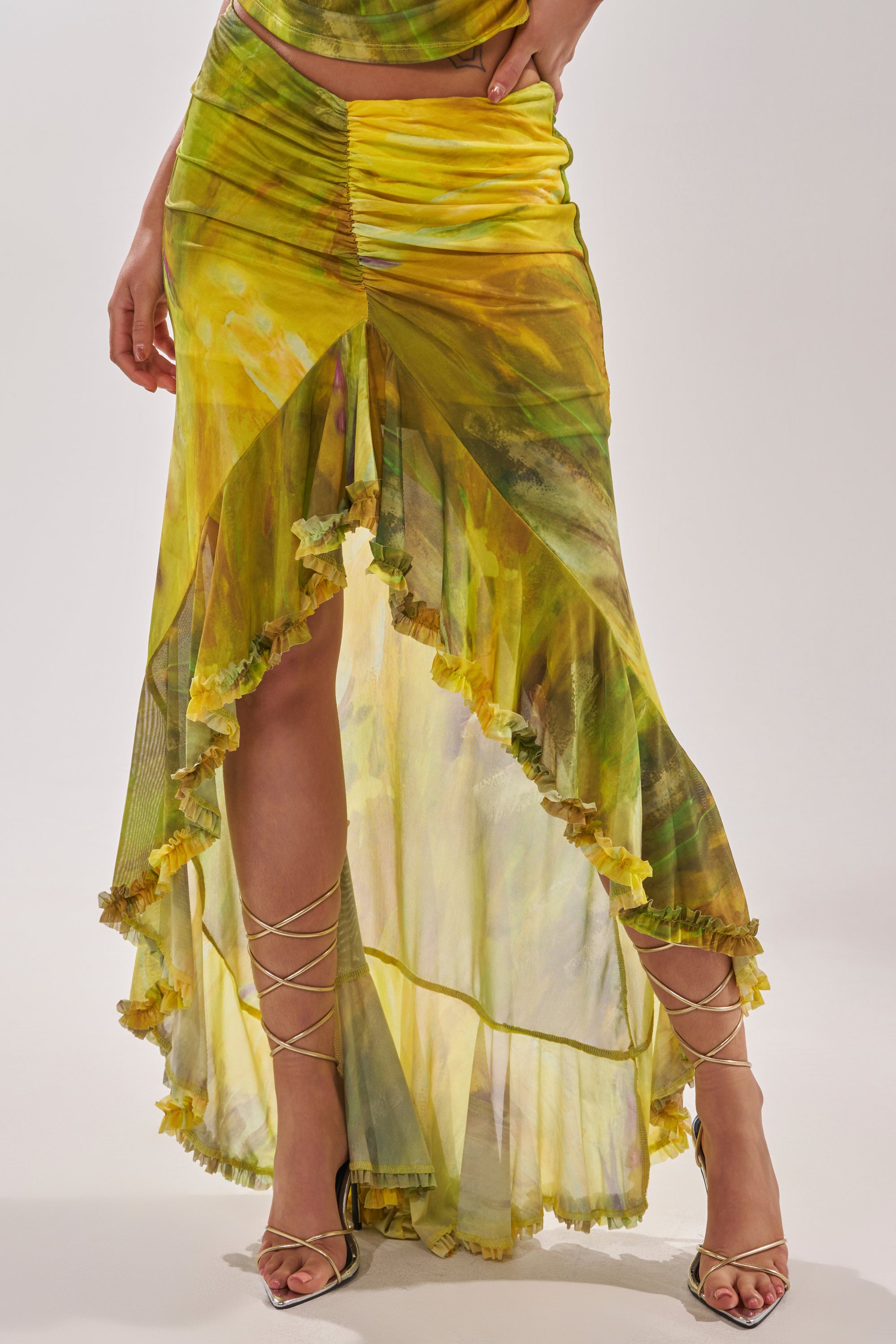A person wearing the TELL ME MORE HIGH LOW SKIRT IN YELLOW, featuring a marble print, ruched detailing, ruffled hem, high slit, and strappy heeled sandals. Only the lower body and part of one hand are visible. 2