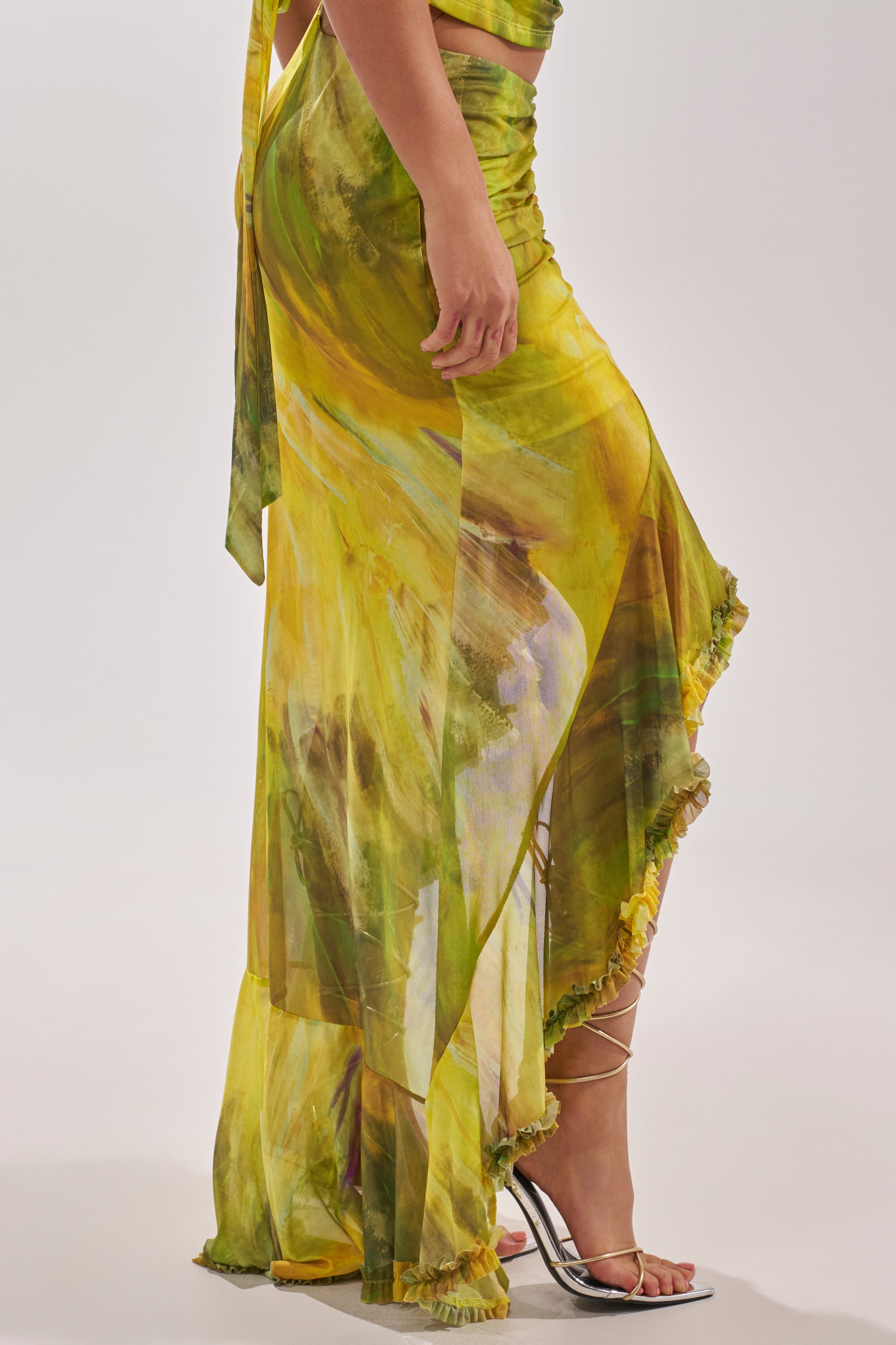 Someone wears the TELL ME MORE HIGH LOW SKIRT IN YELLOW, which features a flowy silhouette, high-low hem, and ruched waist. It's paired with strappy silver heels against a plain, light-colored background. 3