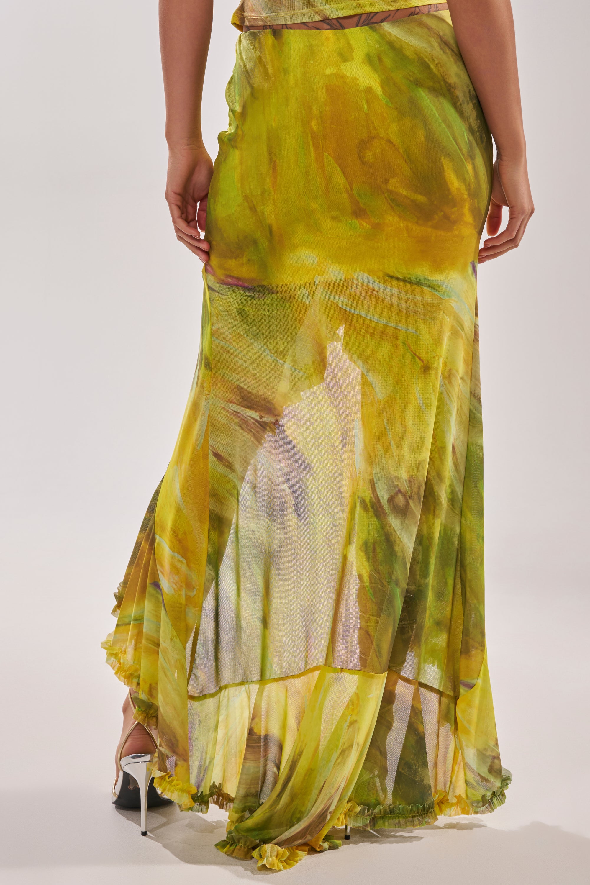 A person wearing the TELL ME MORE HIGH LOW SKIRT IN YELLOW, seen from the back, with high heels visible and the hem brushing the floor. 4