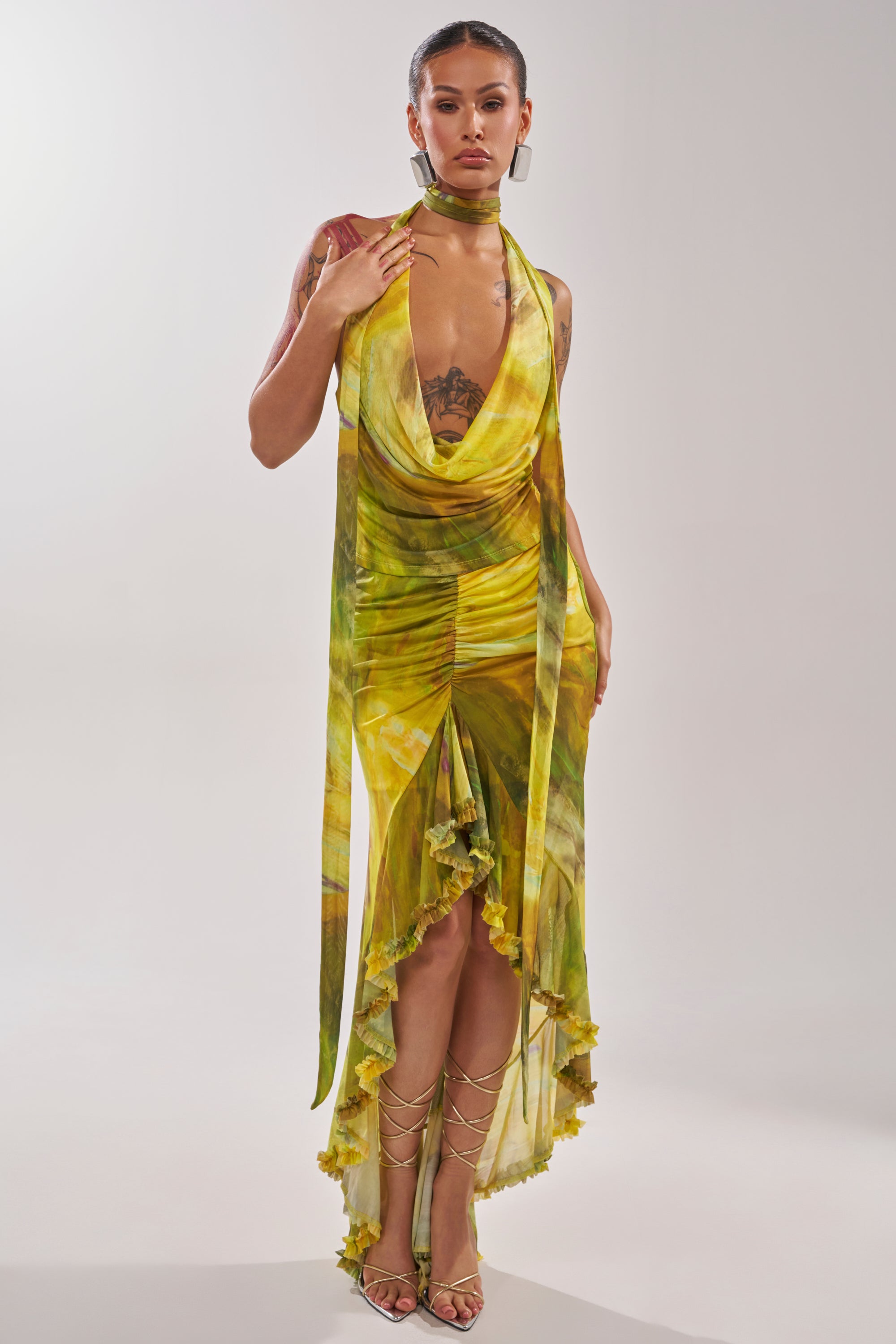A woman showcases the TELL ME MORE HIGH LOW SKIRT IN YELLOW with gold lace-up sandals, hoop earrings, and her tattoos. The skirt features a high-low ruffled hem and a draped design for a bold look. 5