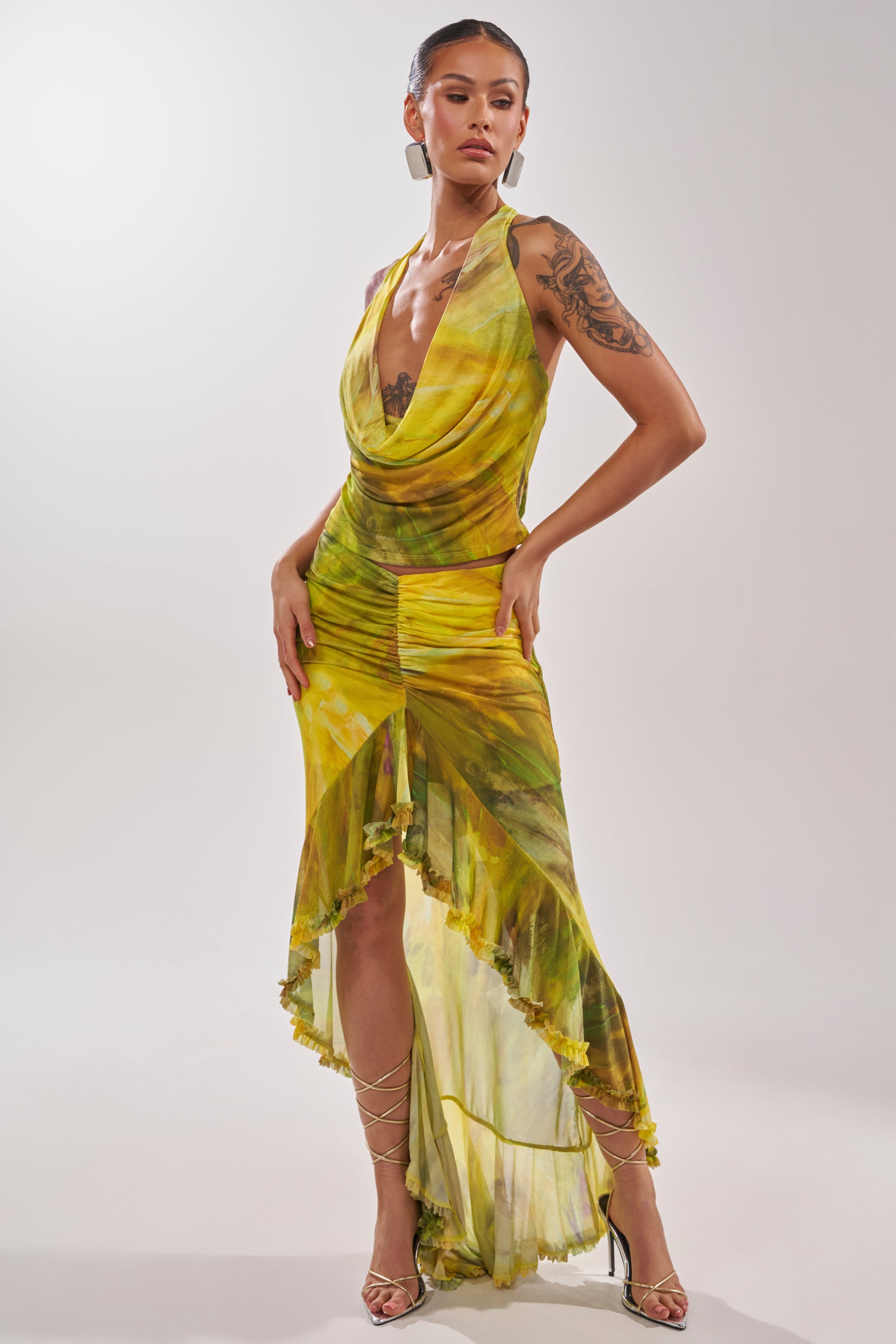 A woman showcases the TELL ME MORE HIGH LOW SKIRT IN YELLOW, styled with strappy silver heels and her hair pulled back to reveal a large arm tattoo. The skirt features a sleeveless yellow and green tie-dye design and draped neckline. 8
