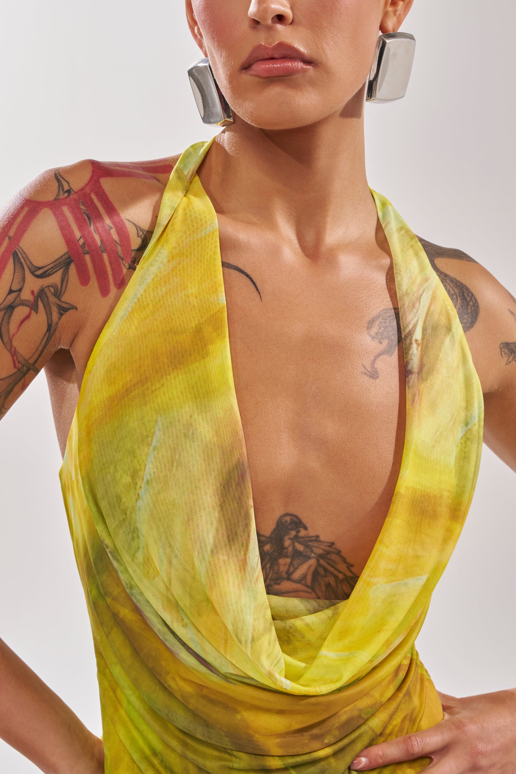 A person wears the TELL ME MORE COWL NECK BLOUSE, a sleeveless yellow-green top with a draped cowl neckline, paired with large silver geometric earrings and vibrant tattoos. A plain, light background highlights their look. 9