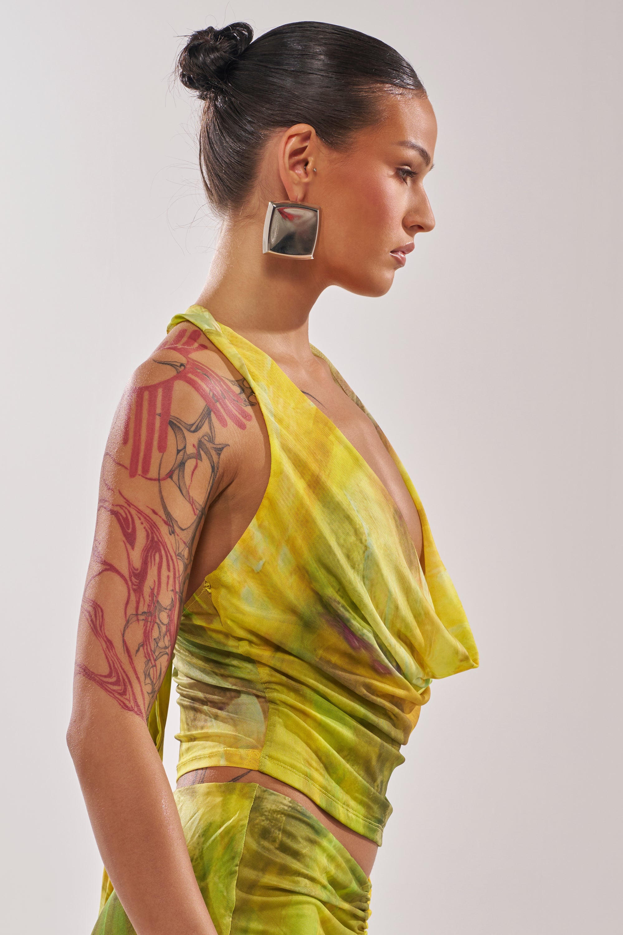 A woman with slicked-back dark hair in a bun, silver square earrings, and a large red abstract arm tattoo stands in profile wearing the TELL ME MORE COWL NECK BLOUSE in yellow-green. 2