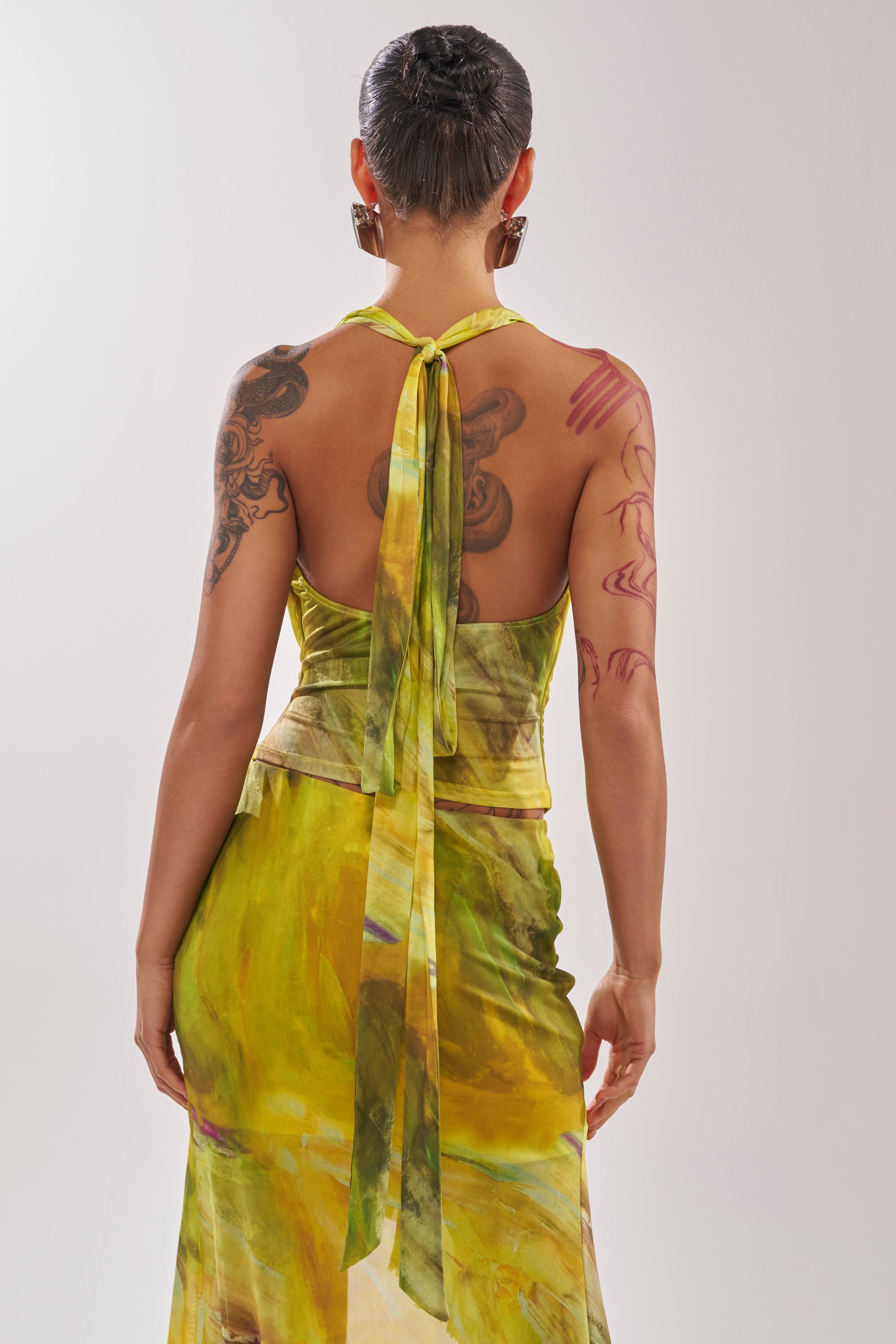 A person with short hair and visible tattoos, shown from behind against a plain light background, wears the TELL ME MORE COWL NECK BLOUSE in yellow-green. 3