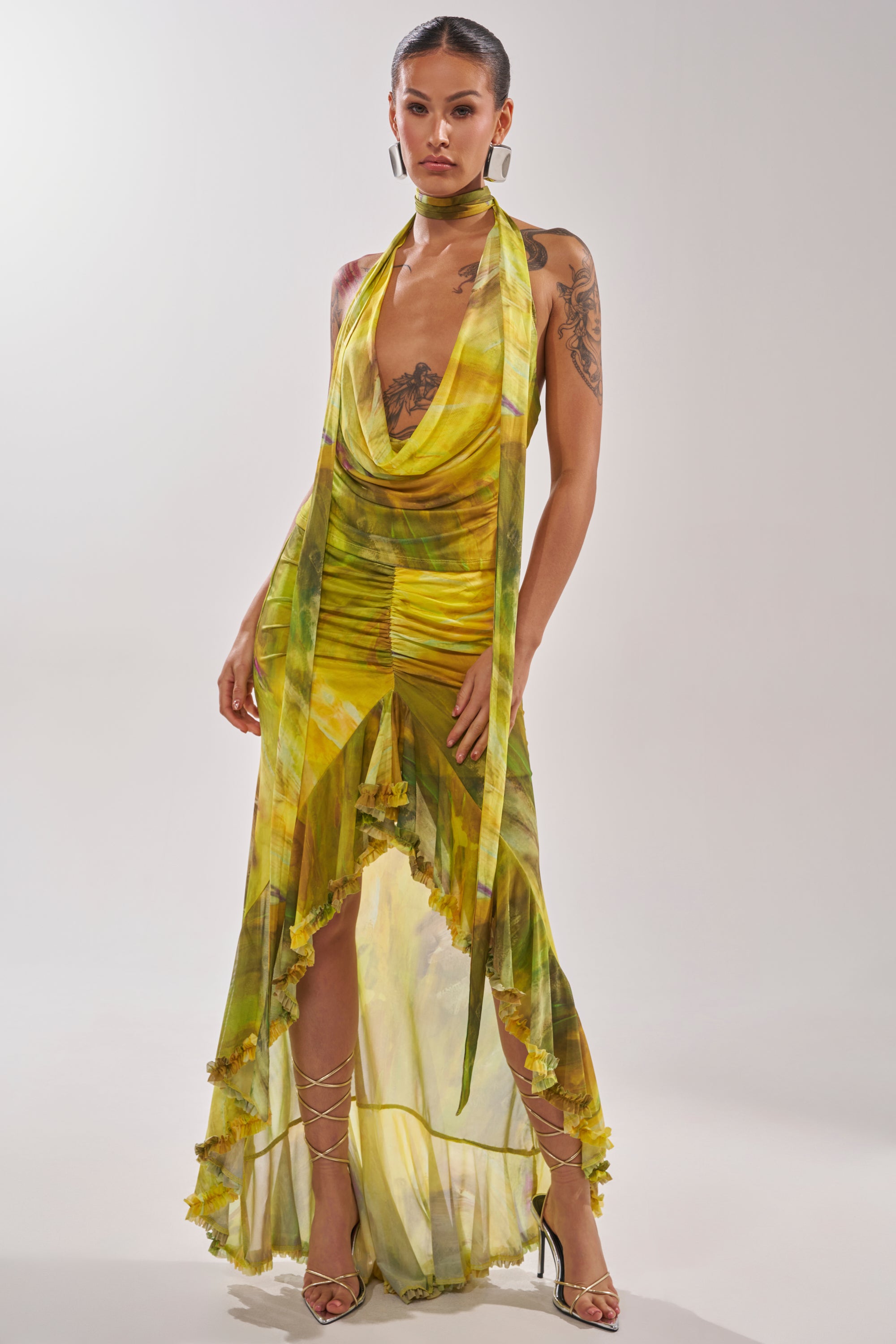 A woman wears the TELL ME MORE COWL NECK BLOUSE in yellow and green marble print with a deep cowl neckline, styled with tattoos, pulled-back hair, and strappy heels as she poses confidently against a plain background. 6