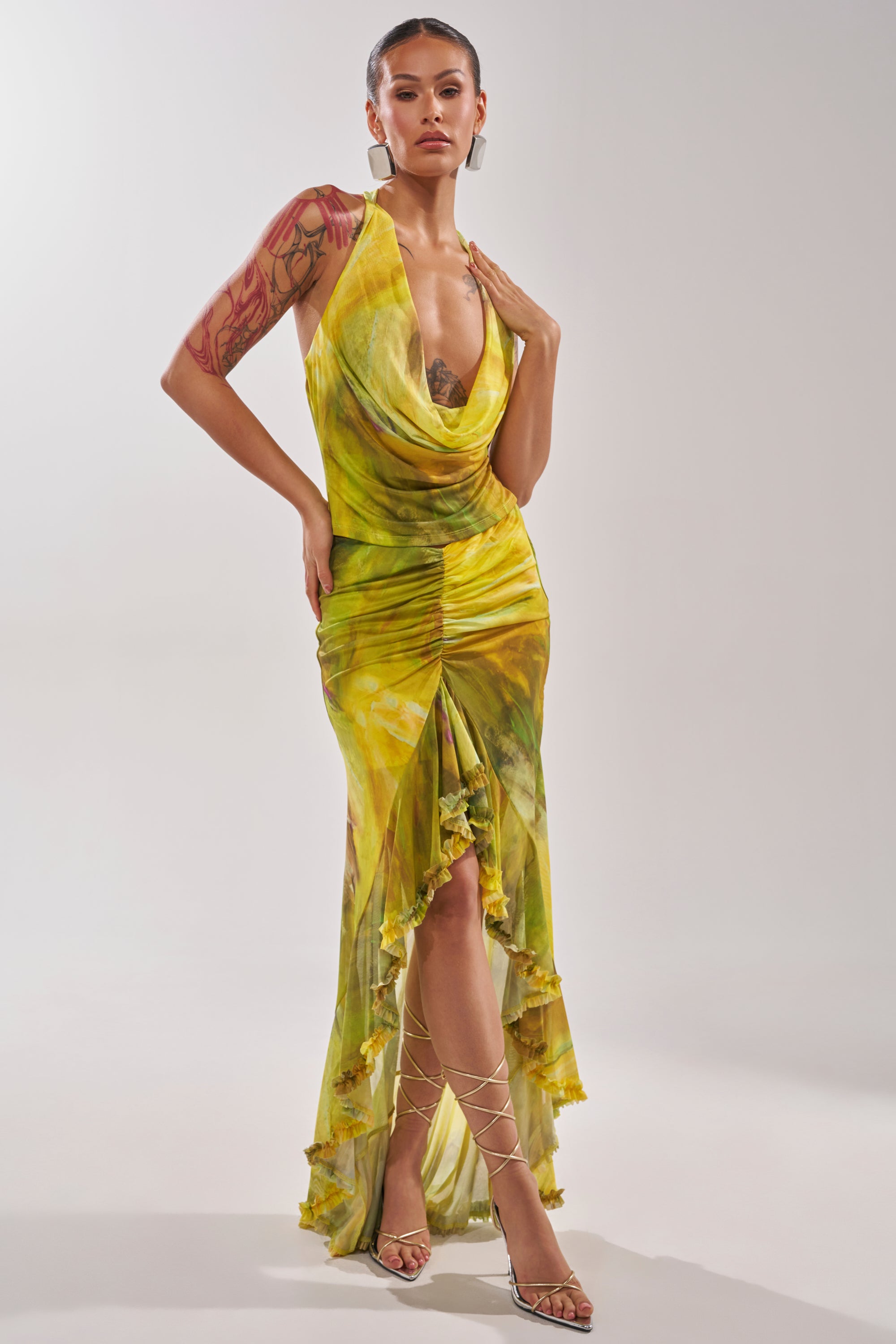 A woman wears the TELL ME MORE COWL NECK BLOUSE in a green marble print, featuring a draped neckline. She has tattoos on her left arm, paired with strappy heels, and stands against a plain light background. 7