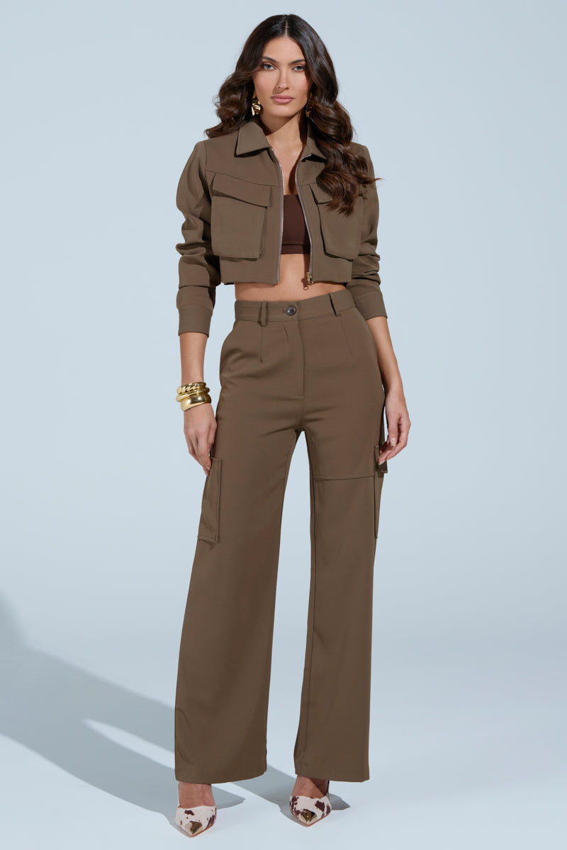 A woman stands against a light grey background wearing the TAKE IT EASY CARGO PANT IN TAUPE, a brown cropped jacket, a dark brown crop top, pointed-toe heels, with long wavy hair and gold bangles.