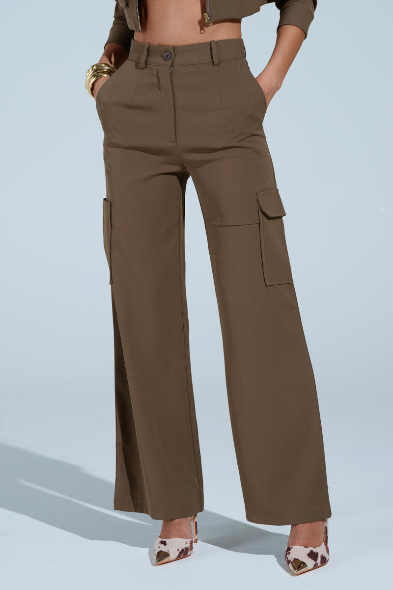 A person wears the TAKE IT EASY CARGO PANT IN TAUPE, styled with a cropped brown jacket and animal print heels. Their hands are tucked in the pants’ side pockets, gold bangles glinting on one wrist.