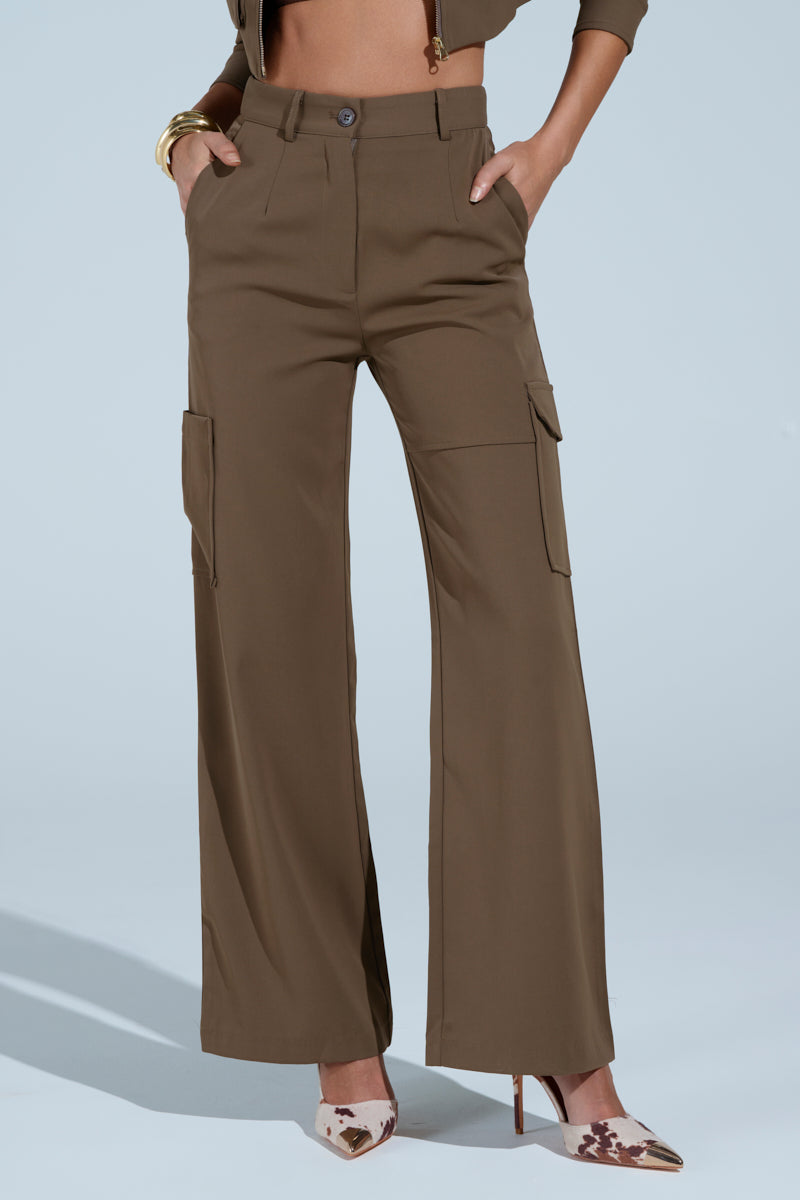 Wearing the TAKE IT EASY CARGO PANT IN TAUPE—high-rise, relaxed straight leg cargo pants with side pockets—paired with brown and white pointed-toe heels. Hands are in pockets; only the lower body is visible.