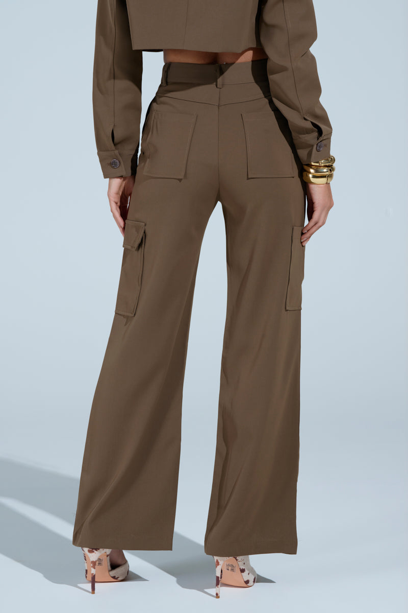 A woman shown from the back wears the TAKE IT EASY CARGO PANT IN TAUPE, high-rise relaxed straight leg style with pockets, paired with a matching cropped jacket, gold bracelets, and patterned high-heel shoes against a plain background.
