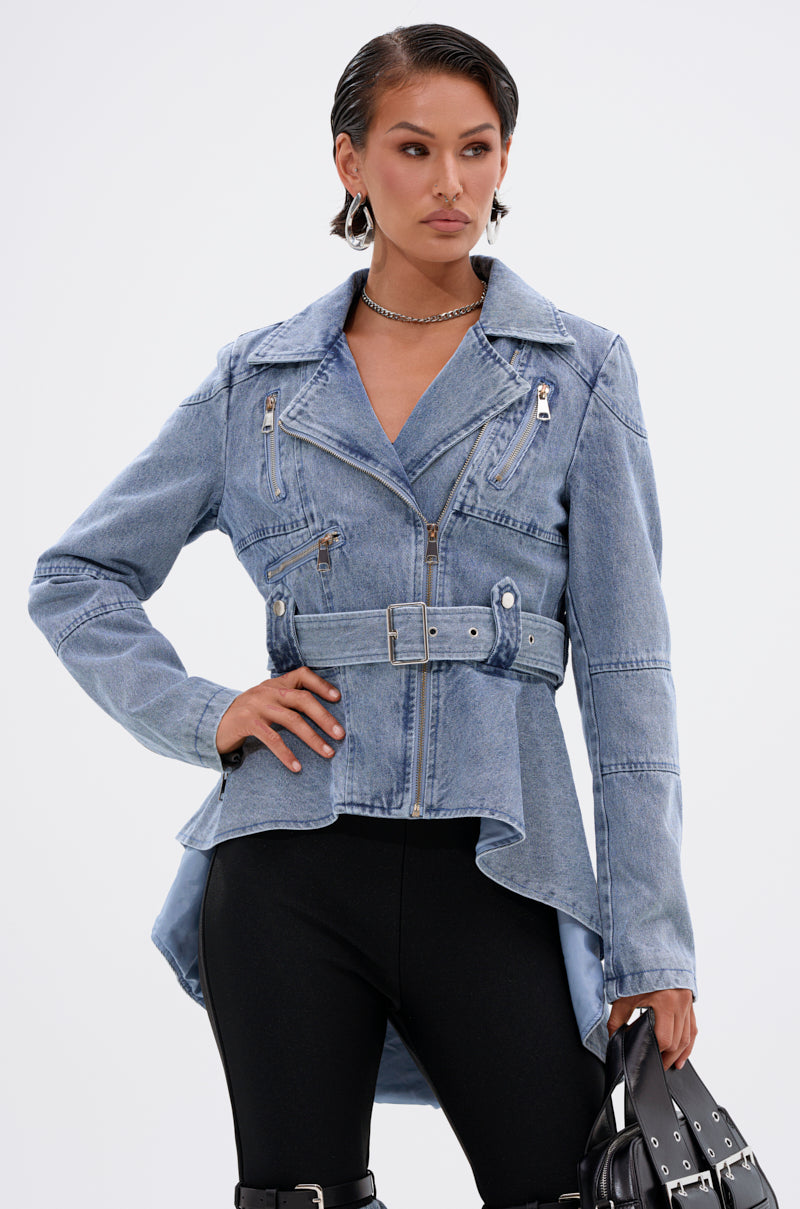 A woman wearing the ASTRO HIGH LOW DENIM JACKET with a belted waist and zip details, paired with black pants, hoop earrings, a silver necklace, and holding a black handbag against a plain white background.
