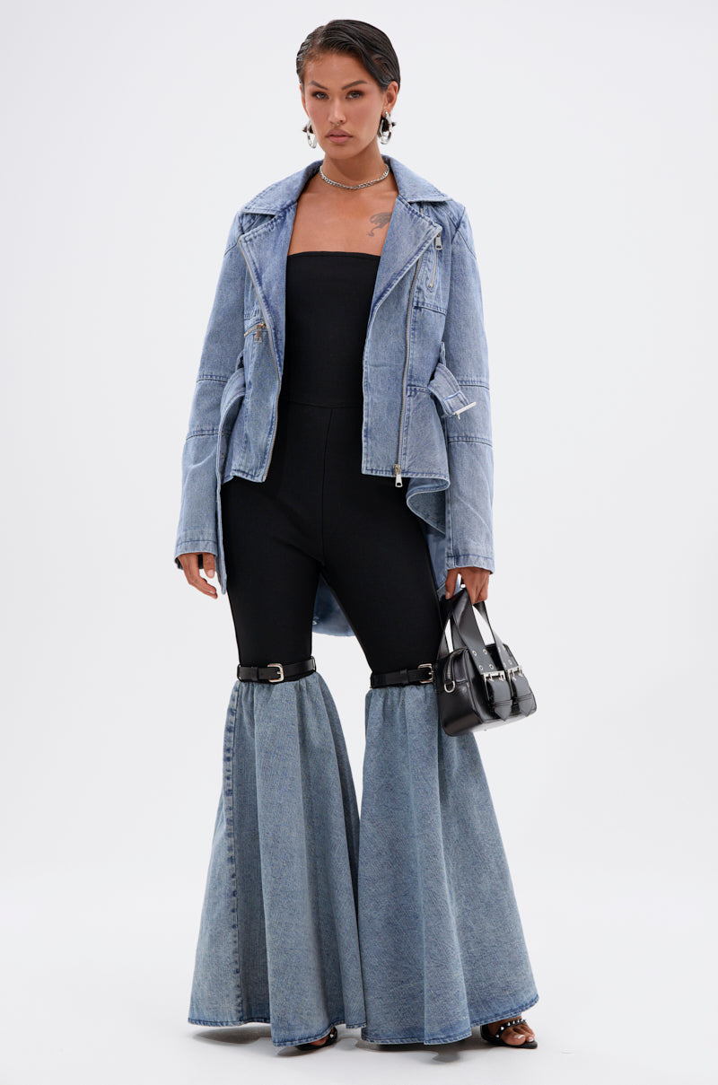 A person poses confidently in the ASTRO HIGH LOW DENIM JACKET, paired with a black strapless top and unique flared black denim pants. They accessorize with a small black handbag and hoop earrings, standing against a plain white background.