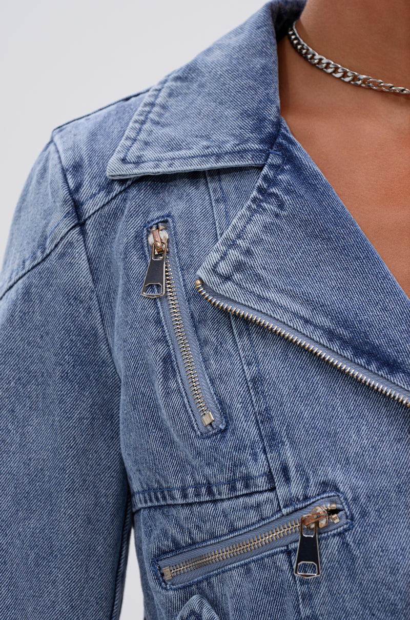 Close-up of a person wearing the ASTRO HIGH LOW DENIM JACKET, a light blue moto-inspired piece with three zippers, a notched collar, textured denim fabric, and styled with a silver chain necklace.