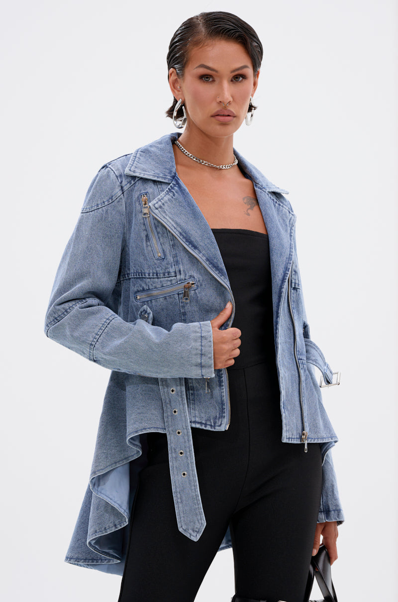 A woman with slicked-back hair wears the ASTRO HIGH LOW DENIM JACKET, featuring a belted waist, over a black strapless top and black pants, styled with silver hoop earrings and a chain necklace against a plain white background.