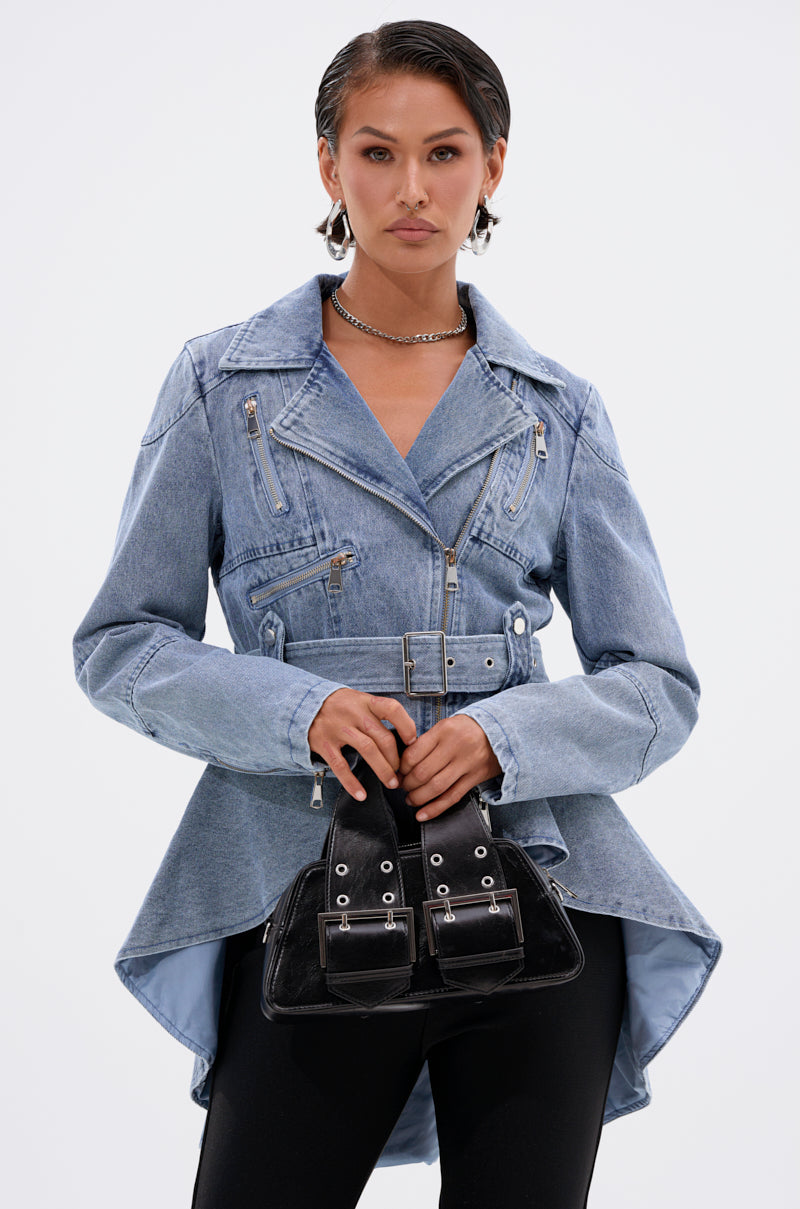 A woman wears the ASTRO HIGH LOW DENIM JACKET in light blue with zipper details and a belted waist, paired with black pants. She holds a black handbag with silver hardware, her slicked-back hair and hoop earrings enhancing her confidence.