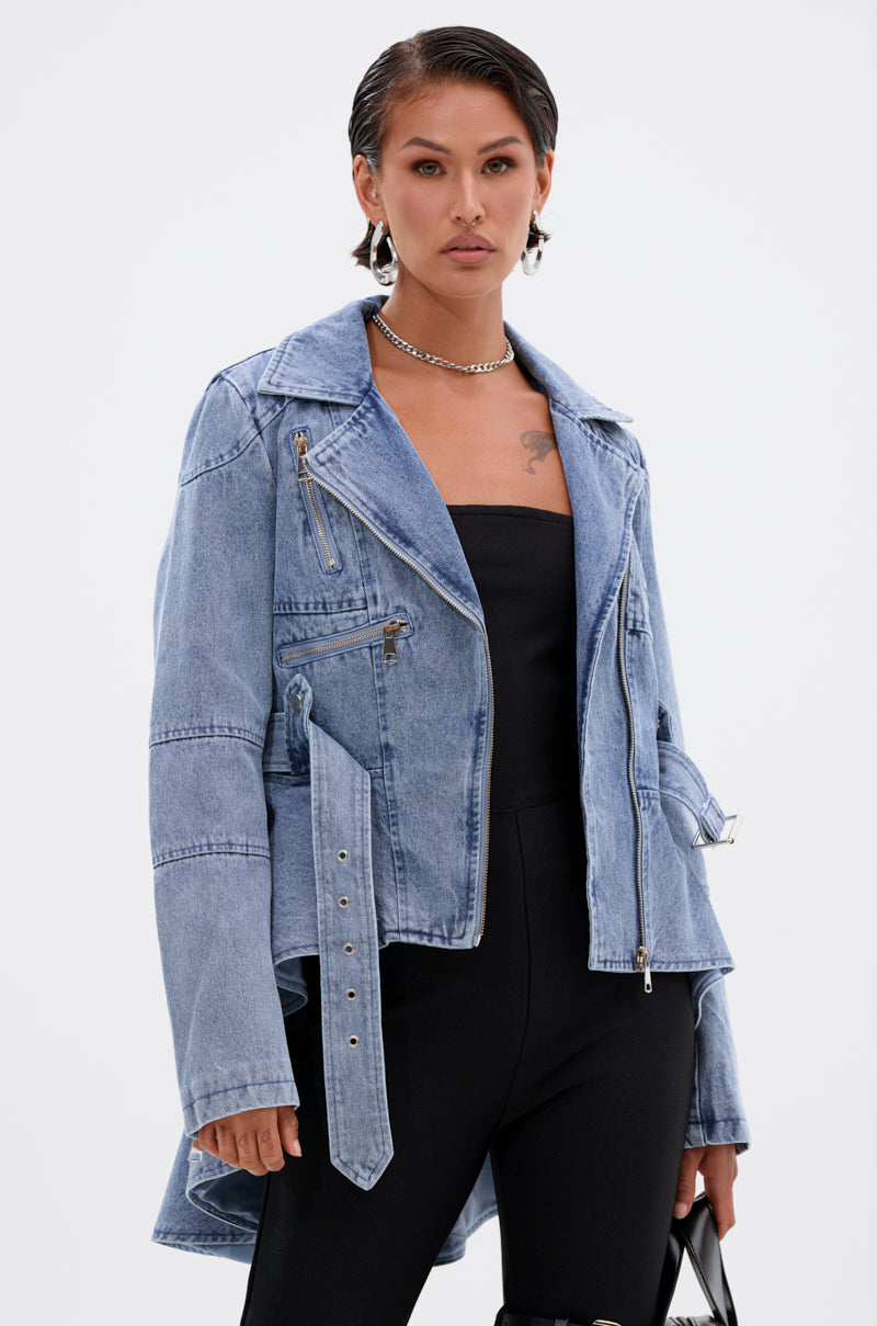 A person with short, slicked-back hair wears silver hoop earrings, a chunky necklace, the ASTRO HIGH LOW DENIM JACKET with zippers and belt details, a black strapless outfit, and holds a small bag.