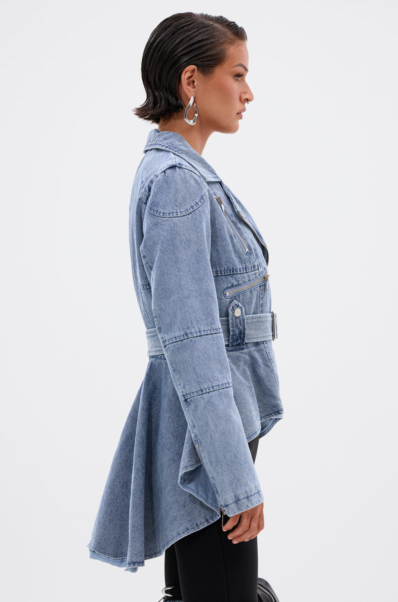 Someone in profile wears the ASTRO HIGH LOW DENIM JACKET in light blue, featuring a belted waist and flared asymmetrical hem, paired with black pants. They have short slicked-back hair and silver hoop earrings against a plain white background.