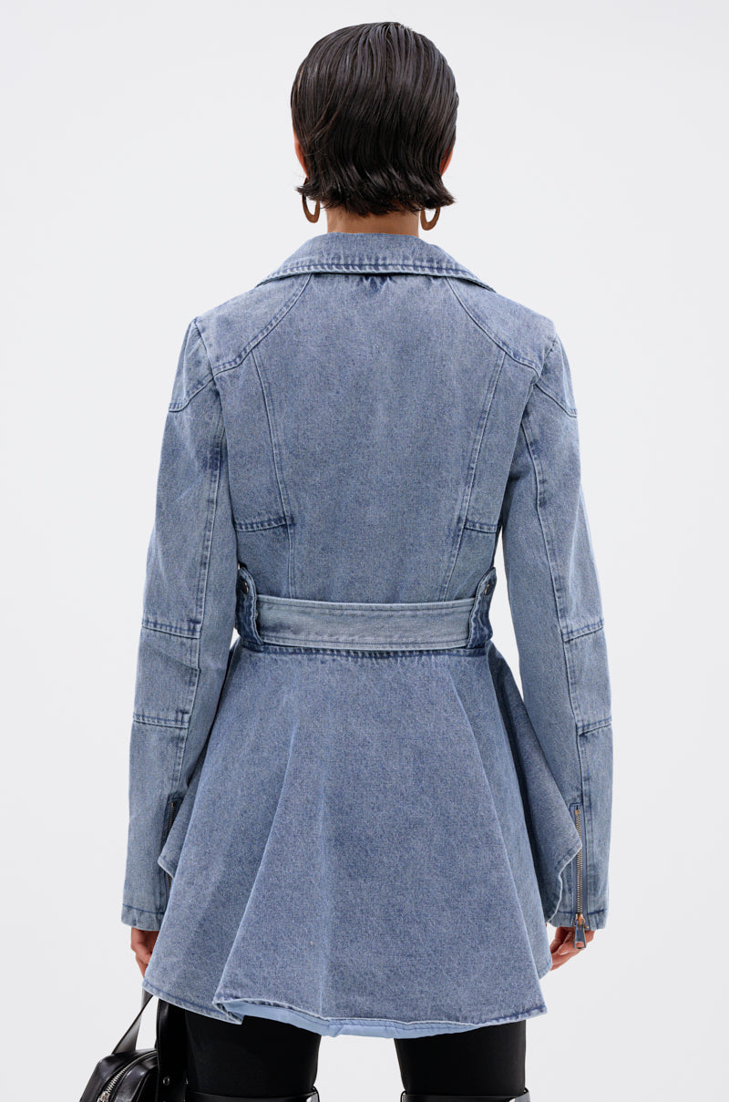 A person with short dark hair, seen from behind, wears the ASTRO HIGH LOW DENIM JACKET with a belted waist and flared hem, holding a black handbag against a plain light background.