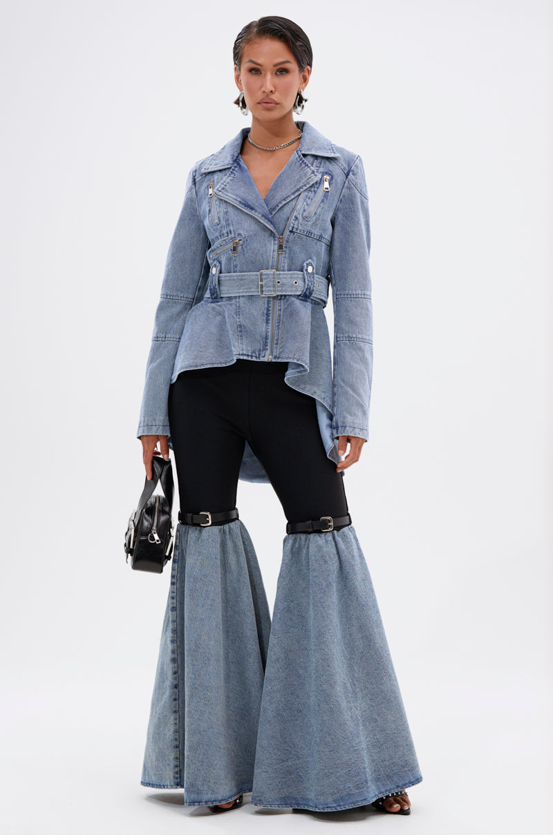 A woman poses against a plain background wearing the ASTRO HIGH LOW DENIM JACKET with black pants and wide-leg denim chaps. She holds a small black handbag and wears statement earrings, creating a bold, modern look.