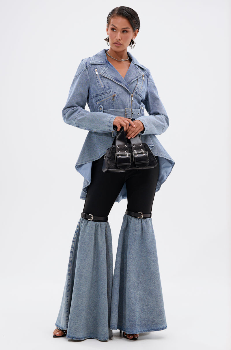 A woman exudes confidence in the ASTRO HIGH LOW DENIM JACKET paired with black flared pants, denim chaps, and a small black handbag. Her short slicked-back hair and hoop earrings enhance her edgy style.