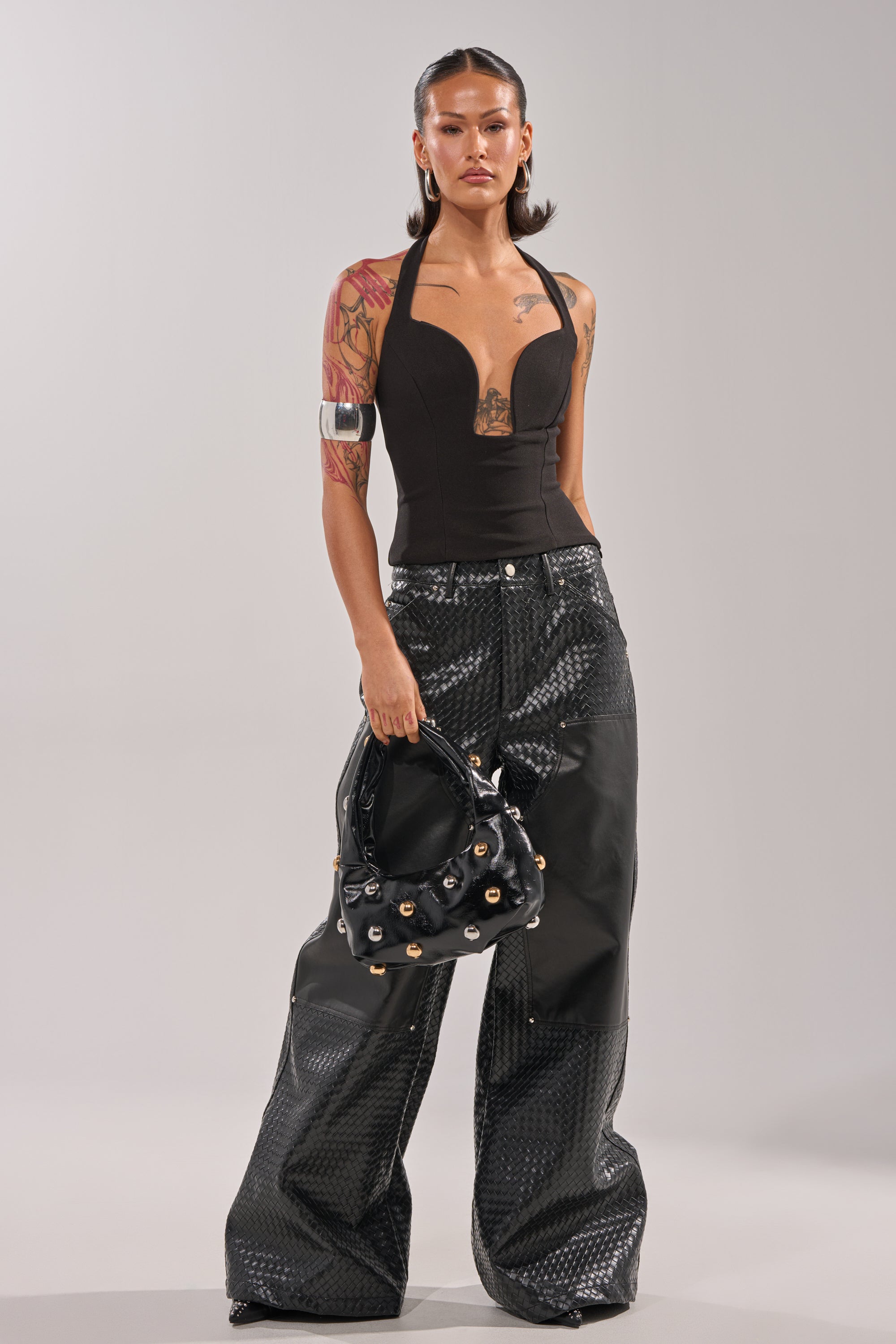 A woman with tattoos wears the TRY ME HALTER TOP IN BLACK and shiny, wide-leg black pants. She holds a black handbag with gold details and has slicked-back hair, standing confidently against a plain background. 4