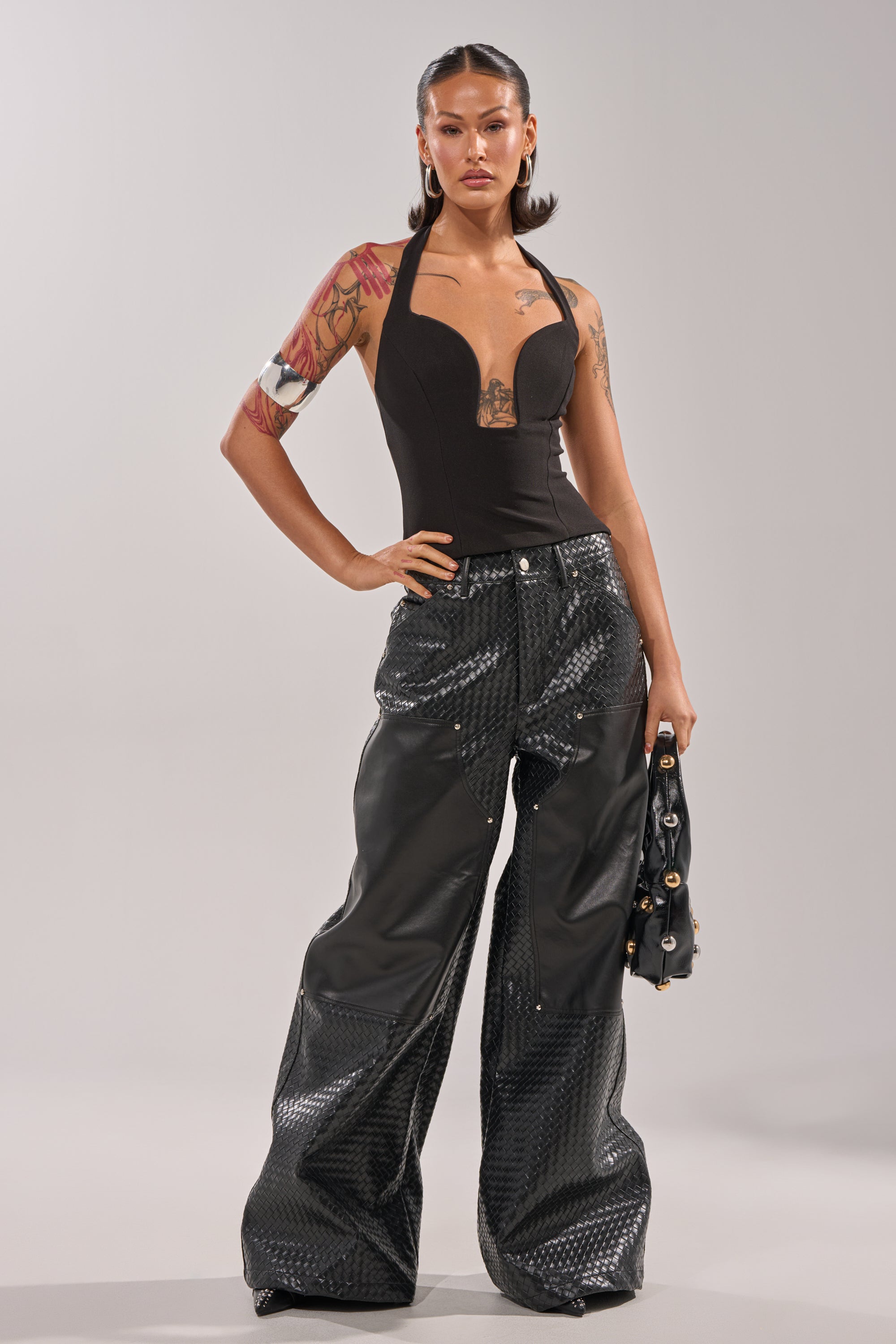 A tattooed woman poses confidently in the TRY ME HALTER TOP IN BLACK with a plunging V-neckline, paired with shiny textured wide-leg pants. She holds a black bag, accessorizing with large hoop earrings and slicked-back hair. 6