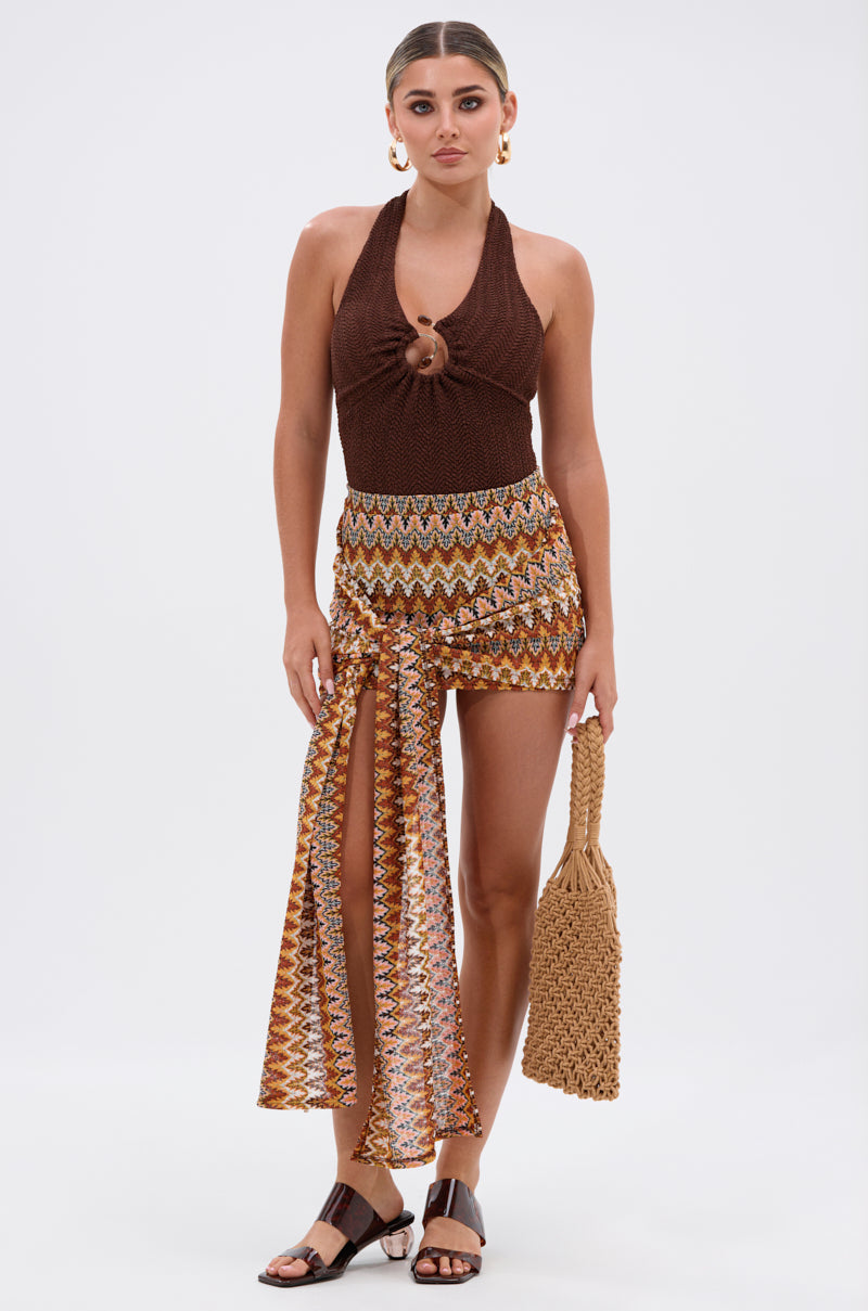 A woman faces forward wearing the GROOVING AND MOVING BODYSUIT styled as a brown halter top, paired with a multicolored zigzag wrap skirt with a high slit, black sandals, gold hoop earrings, and holding a woven tan handbag.