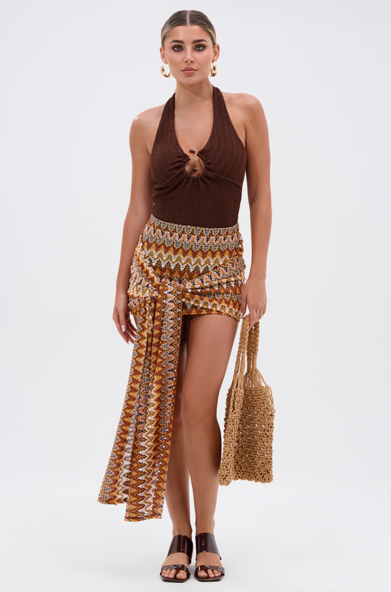 A woman wears the GROOVING AND MOVING BODYSUIT in brown with a patterned asymmetric mini skirt, black sandals, gold hoop earrings, and a woven bag. Her hair is pulled back and the background is plain white.