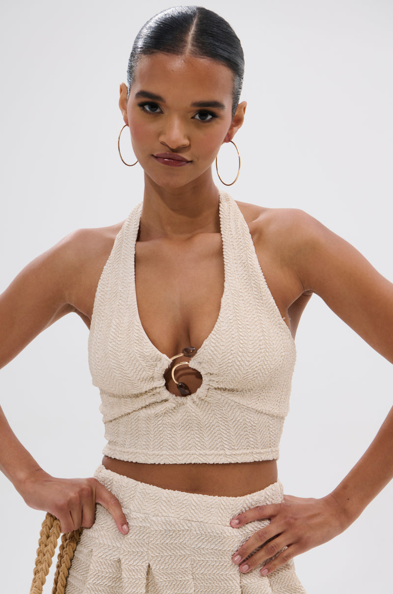 A woman with slicked-back hair and large hoop earrings models the cream HOLLYWOOD SLINKY TEXTURED BRALETTE halter crop top with chest ring detail, paired with matching high-waisted bottoms, posing confidently with one hand on her hip.