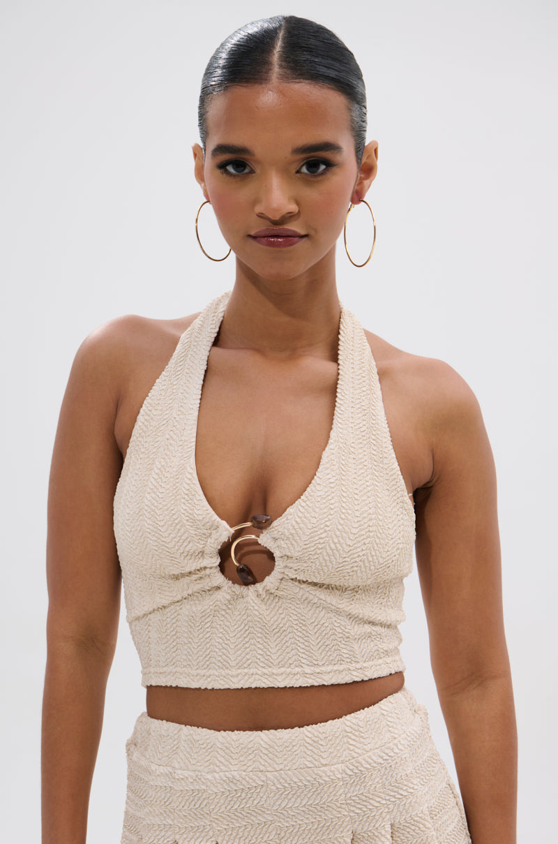 A woman with dark hair in a ponytail and large hoop earrings models the HOLLYWOOD SLINKY TEXTURED BRALETTE, featuring a halter neckline and central ring detail, against a plain white background for a boho-inspired look.