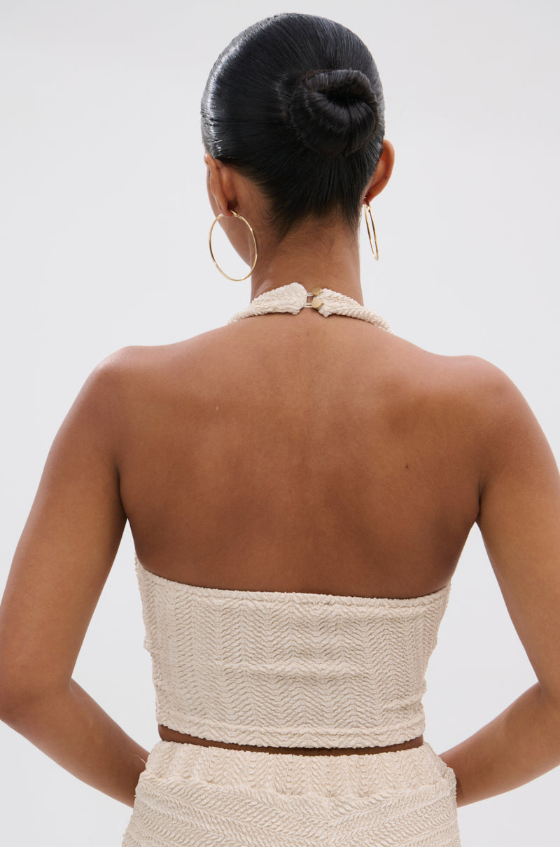 A woman with sleek dark hair in a bun is seen from behind, wearing large hoop earrings and the cream HOLLYWOOD SLINKY TEXTURED BRALETTE with a matching halter strap, lending subtle boho flair to her look.