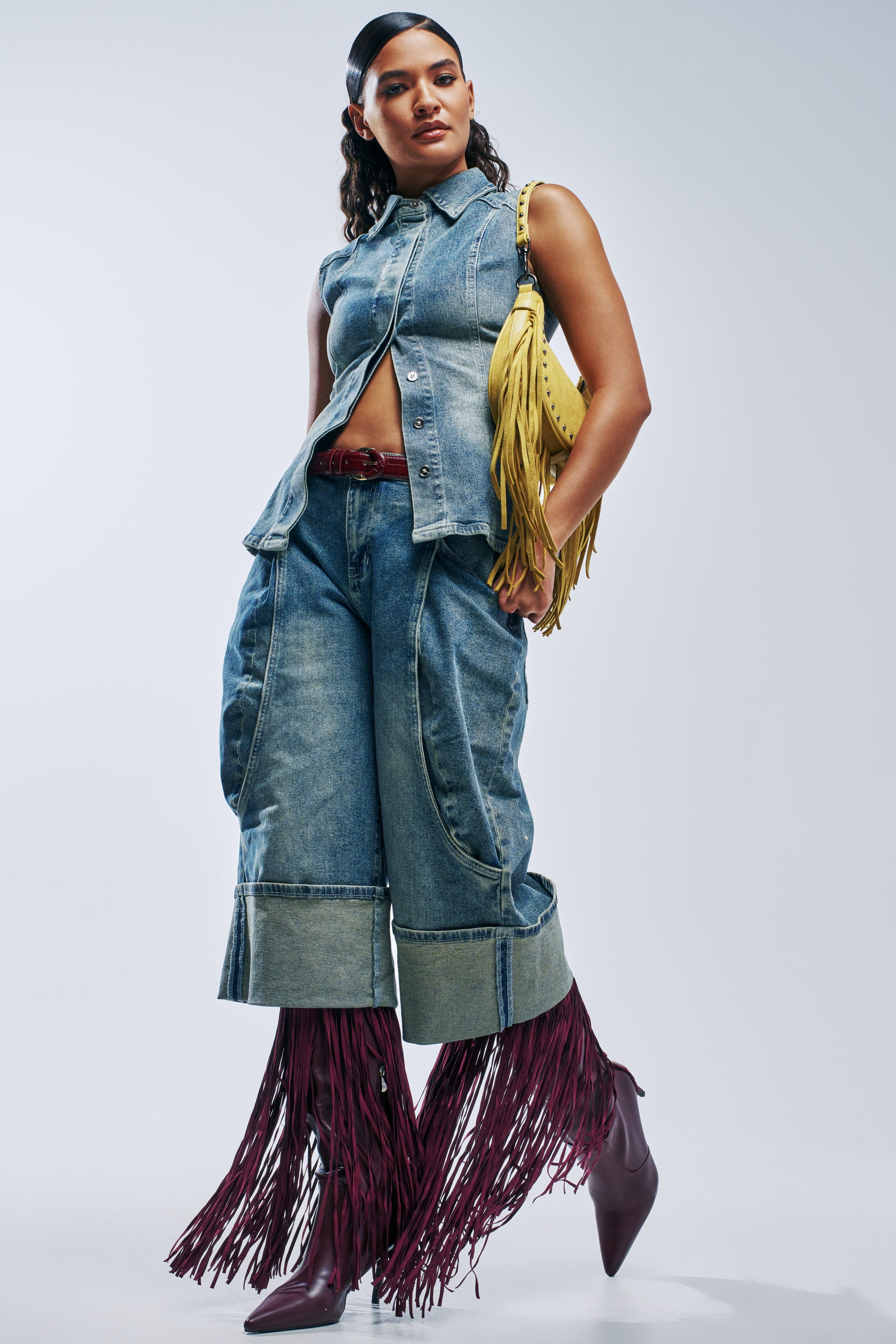A model wearing a sleeveless denim vest and AUBERGINE DENIM CULOTTE, styled with a yellow fringe handbag and burgundy boots featuring long fringe accents.