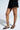Someone in a black skirt is wearing the AKIRA AULORA Black Flat Sandal with Peep Toe, featuring thin vegan leather crisscrossing ankle straps and a cushioned insole for comfort, set against a light, neutral backdrop.