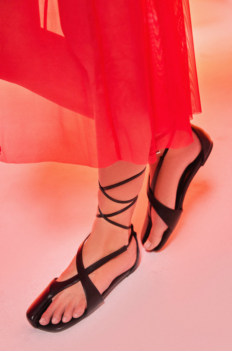 A person stands on a light pink surface, partially covered by a sheer red dress, wearing AKIRA AULORA BLACK FLAT SANDALS WITH PEEP TOE featuring toe loops and ankle ties.