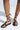 Bare feet in AKIRA AULORA Black Flat Sandals with an open square toe and unique design. These vegan leather sandals have long straps crisscrossing the ankles, a cushioned insole for comfort, all set against a neutral, well-lit background.