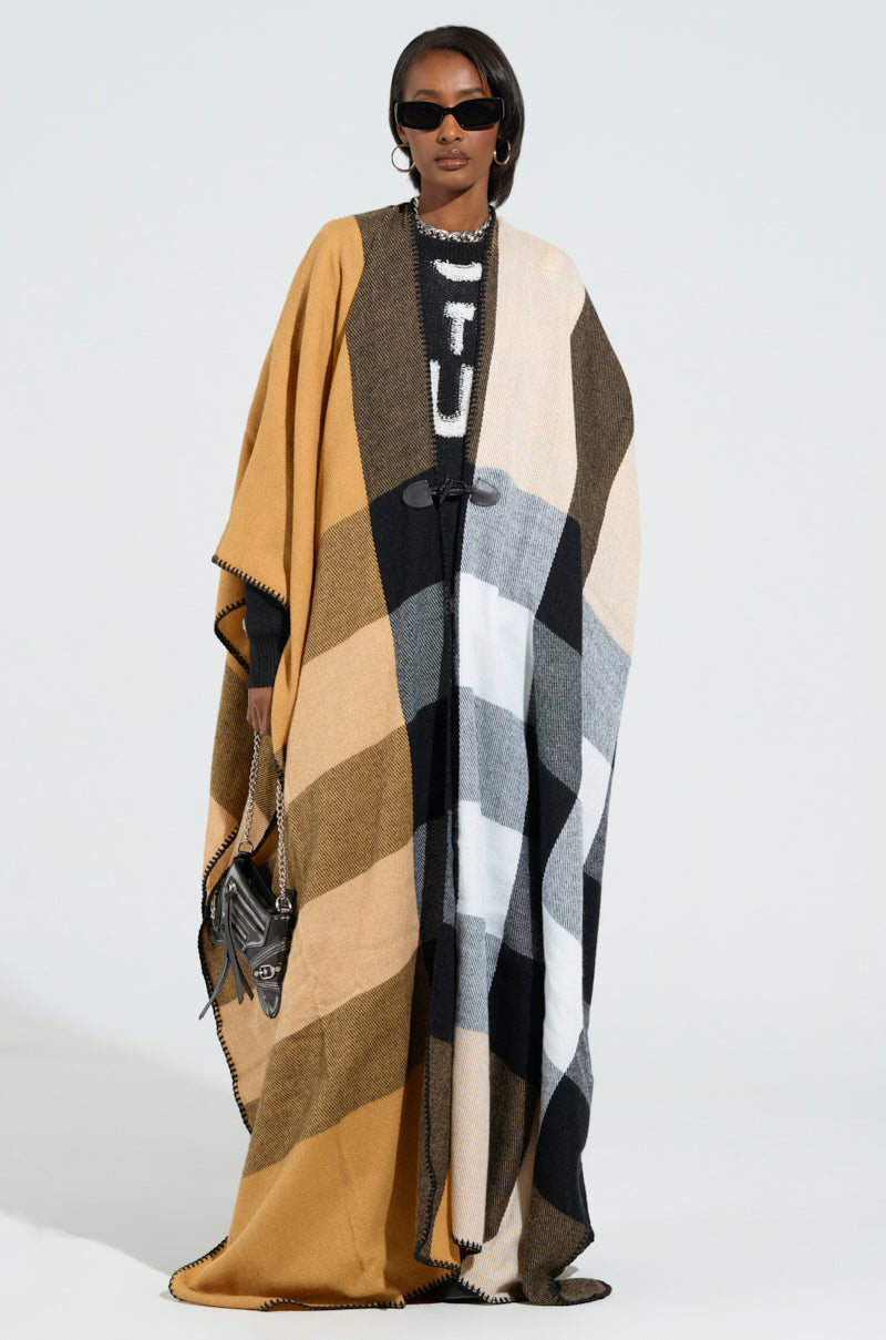 Against a plain backdrop, a person wearing sunglasses showcases the AURETA NEW PLAID MAXI PONCHO, an ultra-soft knit cloak designed with multicolored patchwork in shades of brown, gray, black, and white. The outfit is completed with a horn toggle button and complemented by a handbag. 0