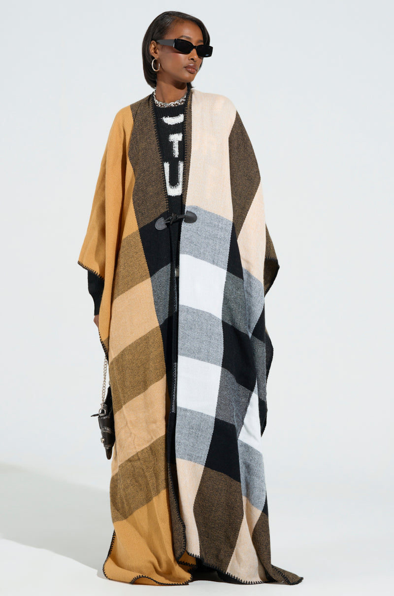 Dressed in the AURETA NEW PLAID MAXI PONCHO, a person exudes confidence while wearing stylish sunglasses. The poncho, showcasing hues of yellow, brown, black, and white in ultra-soft knit fabric, complements their look as they hold a small bag secured with an eye-catching horn toggle button. The background features a gentle, light color palette. 1