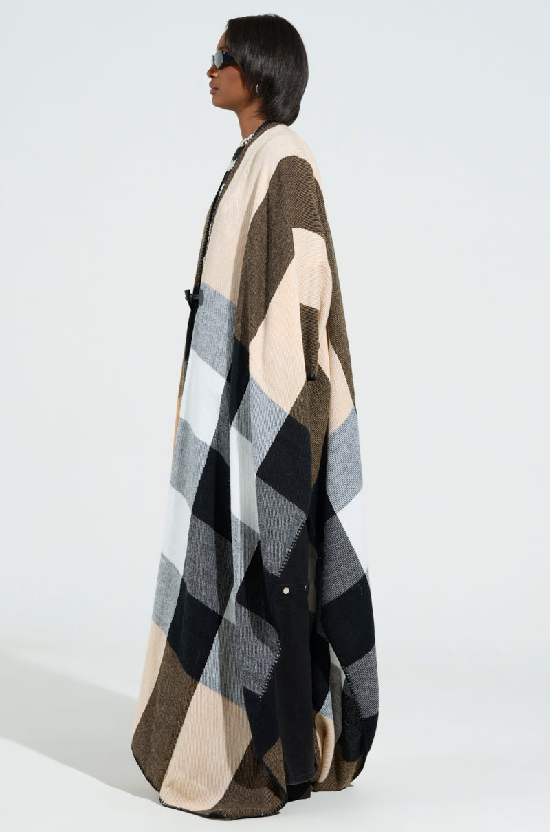 A person with shoulder-length hair is wearing dark sunglasses and the AURETA NEW PLAID MAXI PONCHO, adorned with horn toggle buttons. The ultra-soft knit fabric drapes gracefully in shades of beige, black, and white as they stand in profile against a neutral background. 2