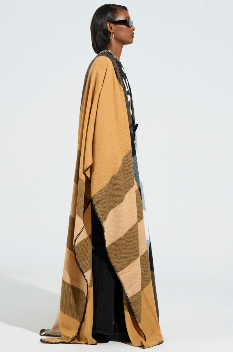 A person wearing sunglasses and the AURETA NEW PLAID MAXI PONCHO, with its elegant, flowing design in shades of brown, black, and camel. The ultra-soft knit fabric drapes gracefully as they stand in profile against a light gray background, adding a chic and sophisticated look to the scene. 3