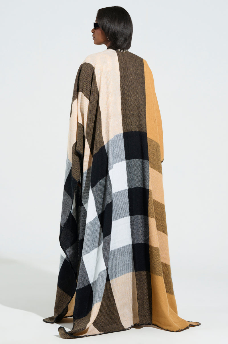A person with shoulder-length hair, wearing sunglasses, is wrapped in the Aureta New Plaid Maxi Poncho, an ultra-soft knit featuring a checkered pattern in shades of black, gray, brown, and cream. They stand against a plain background with the horn toggle button fastened securely. 4