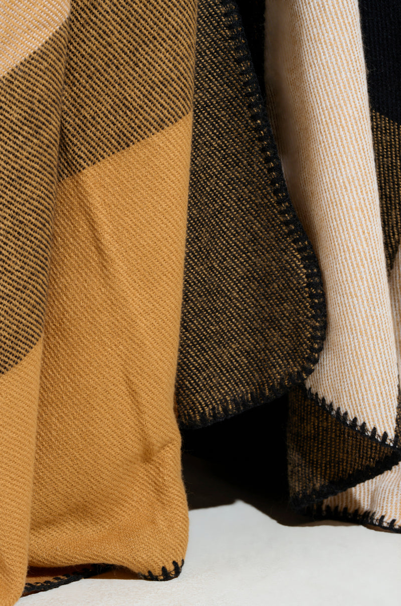 A close-up of the ultra-soft knit fabric from the AURETA NEW PLAID MAXI PONCHO showcases a striped pattern in shades of tan, black, and cream. The visible weave texture and black stitching along the edges enhance its layered appearance, capturing the elegance reminiscent of a plaid maxi poncho. 6