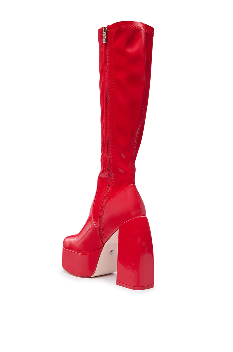 The AZALEA WANG AURON RED LIQUID LUXE PLATFORM BOOT is a shiny, red, knee-high faux patent leather boot with a chunky high heel and side zipper, displayed against a white background.
