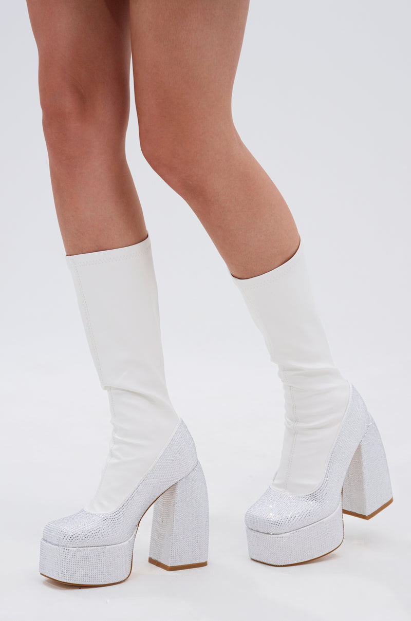 A person wearing the AZALEA WANG AURONNE WHITE AND RHINESTONE CHUNKY BOOT, featuring a glittery platform sole and faux leather finish, stands on a white background.