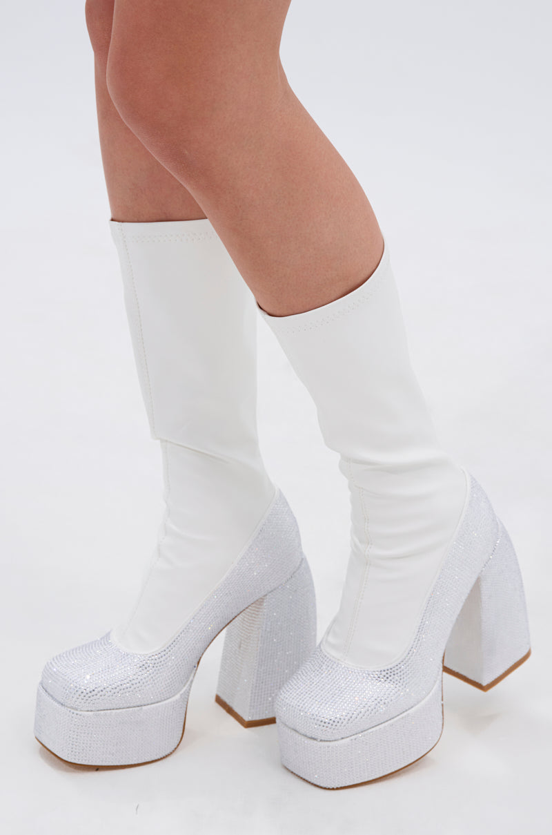 A person stands on a white surface wearing the AZALEA WANG AURONNE WHITE AND RHINESTONE CHUNKY BOOT, featuring a faux leather finish, thick high platform soles with silver glitter, and rhinestone details.
