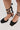 A close-up of lower legs wearing the AZALEA WANG AURORIA BLACK BALLET PLATFORM PUMP with ribbon ankle ties, standing on a light-colored surface.