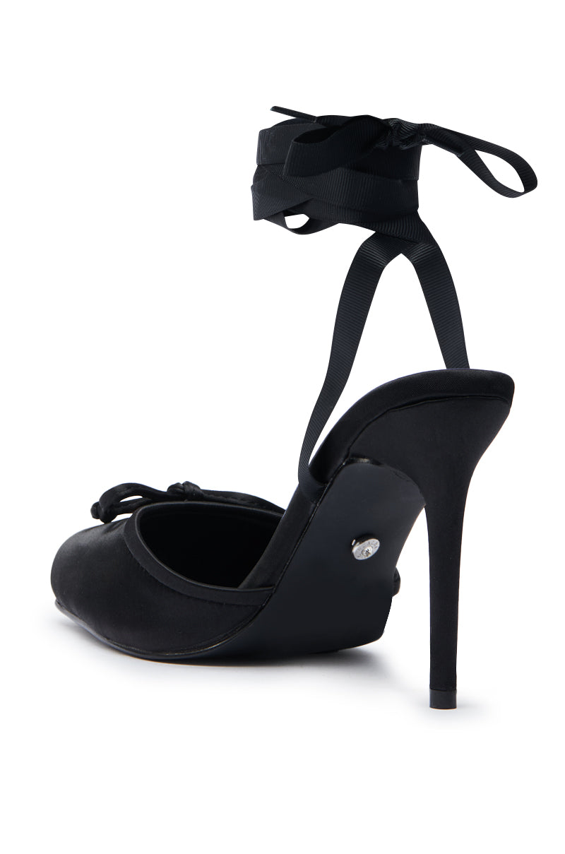 The AZALEA WANG AURORIA BLACK BALLET PLATFORM PUMP features a black satin upper, stiletto heel, and ribbon ankle strap tied in a bow at the back, shown from a rear angle on a white background.