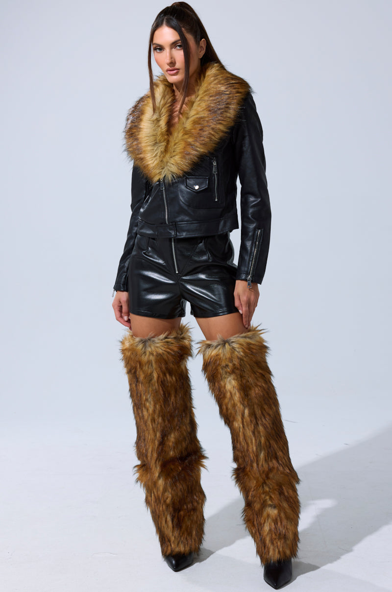 A woman faces forward, wearing the AUSSY FUR COLLAR MOTO black faux leather jacket with a large brown fur collar, paired with black leather shorts and tall brown faux fur leg warmers over black boots, set against a plain light background.